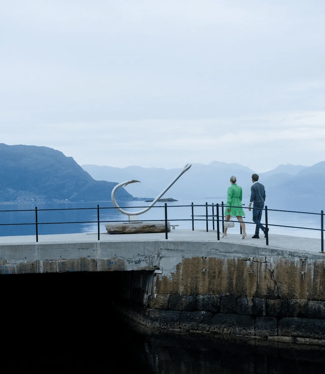 To gjester går langs kaien forbi krok-skulpturen, med fjorden og fjellene bak