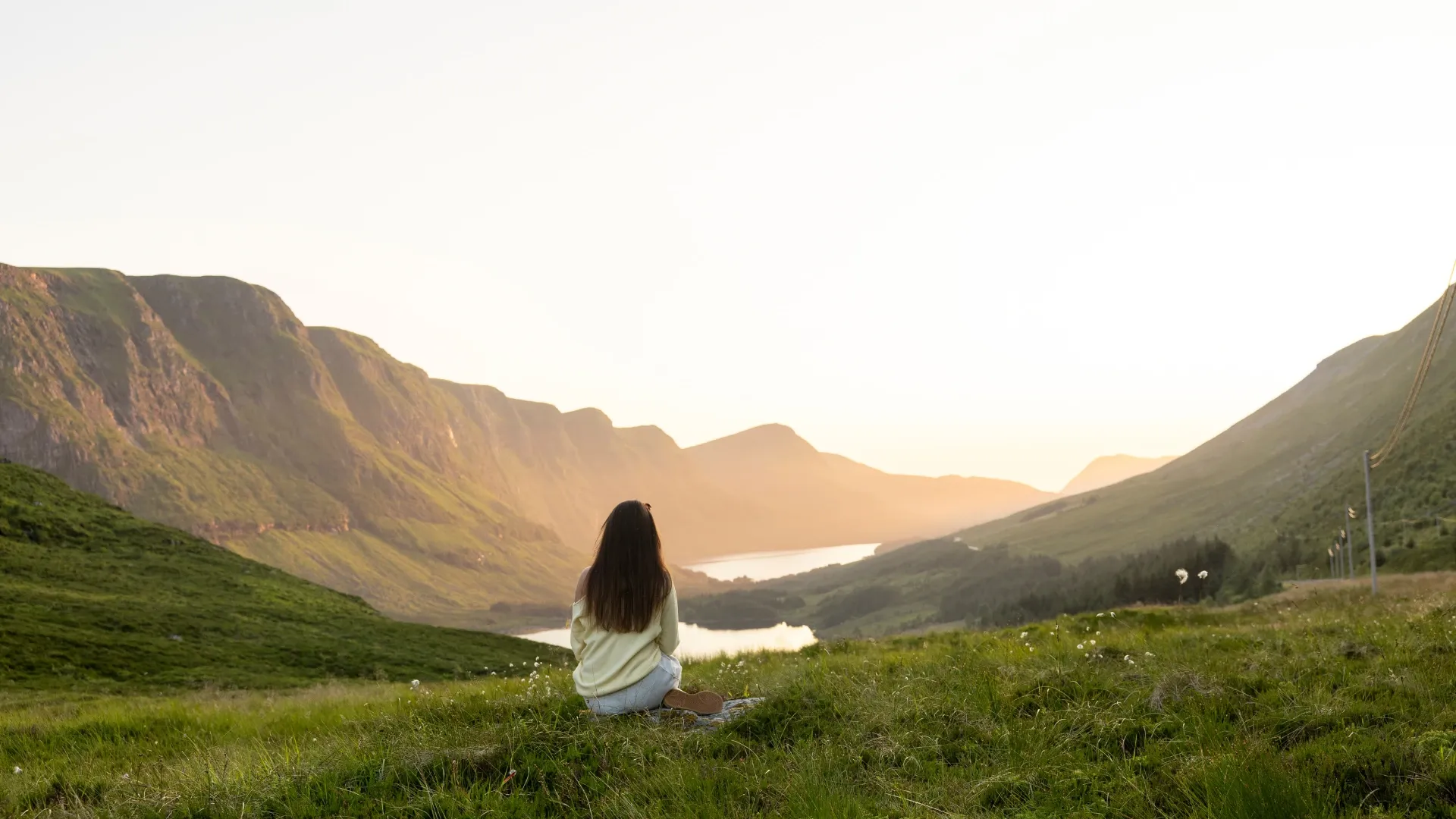 Kvinne sitter i gresset og ser utover dalen i solnedgang